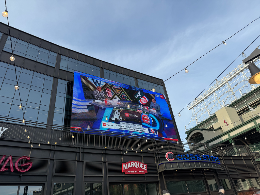 An exterior view of Gallagher Way adjacent to Wrigley Field, looking up at a large outdoor video screen mounted on a modern dark building. The screen broadcasts a Marquee Sports Network pre-game show featuring studio analysts. Overhead string lights crisscross against a clear sky, with the historic green steel structure of Wrigley Field visible to the right.