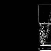 Water being poured into a tall, clear rectangular glass positioned on the right side against a solid black background. Small air bubbles swirl near the bottom left of the liquid inside the glass.