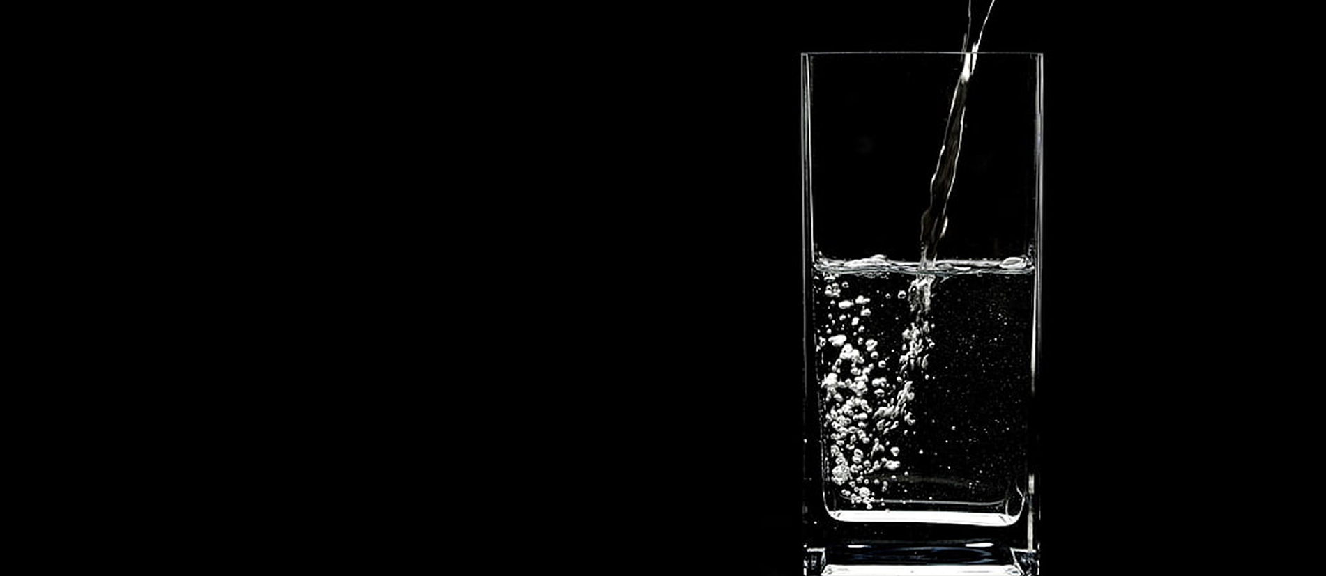Water being poured into a tall, clear rectangular glass positioned on the right side against a solid black background. Small air bubbles swirl near the bottom left of the liquid inside the glass.
