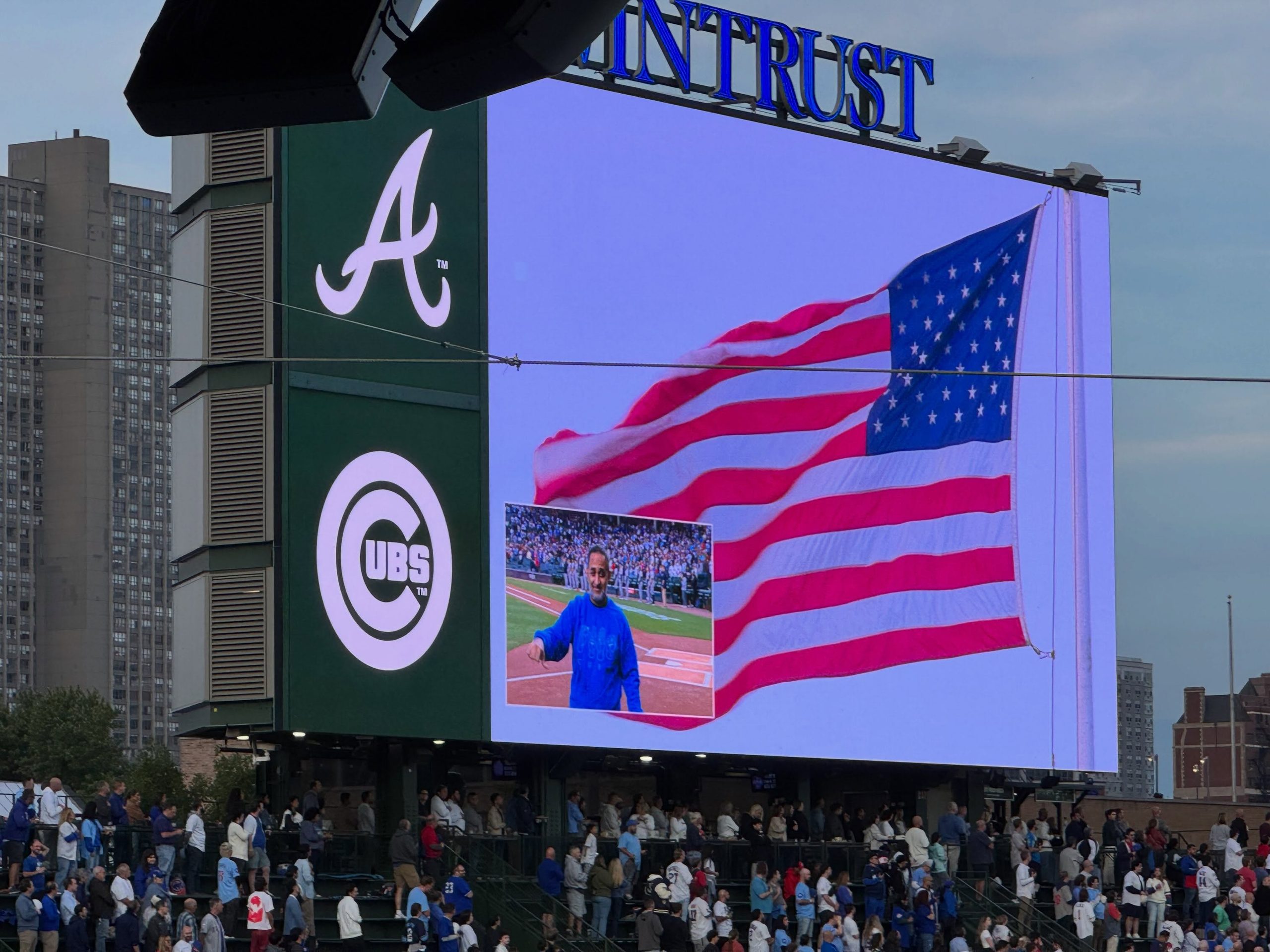 A close-up shot of the large left-field video board at Wrigley Field during pre-game ceremonies. The screen displays an animated American flag alongside a live feed of a performer singing in front of the dugout, framed by the Atlanta Braves "A" logo and the Chicago Cubs circular logo on the left panel. Fans are gathered in the bleacher seats below.