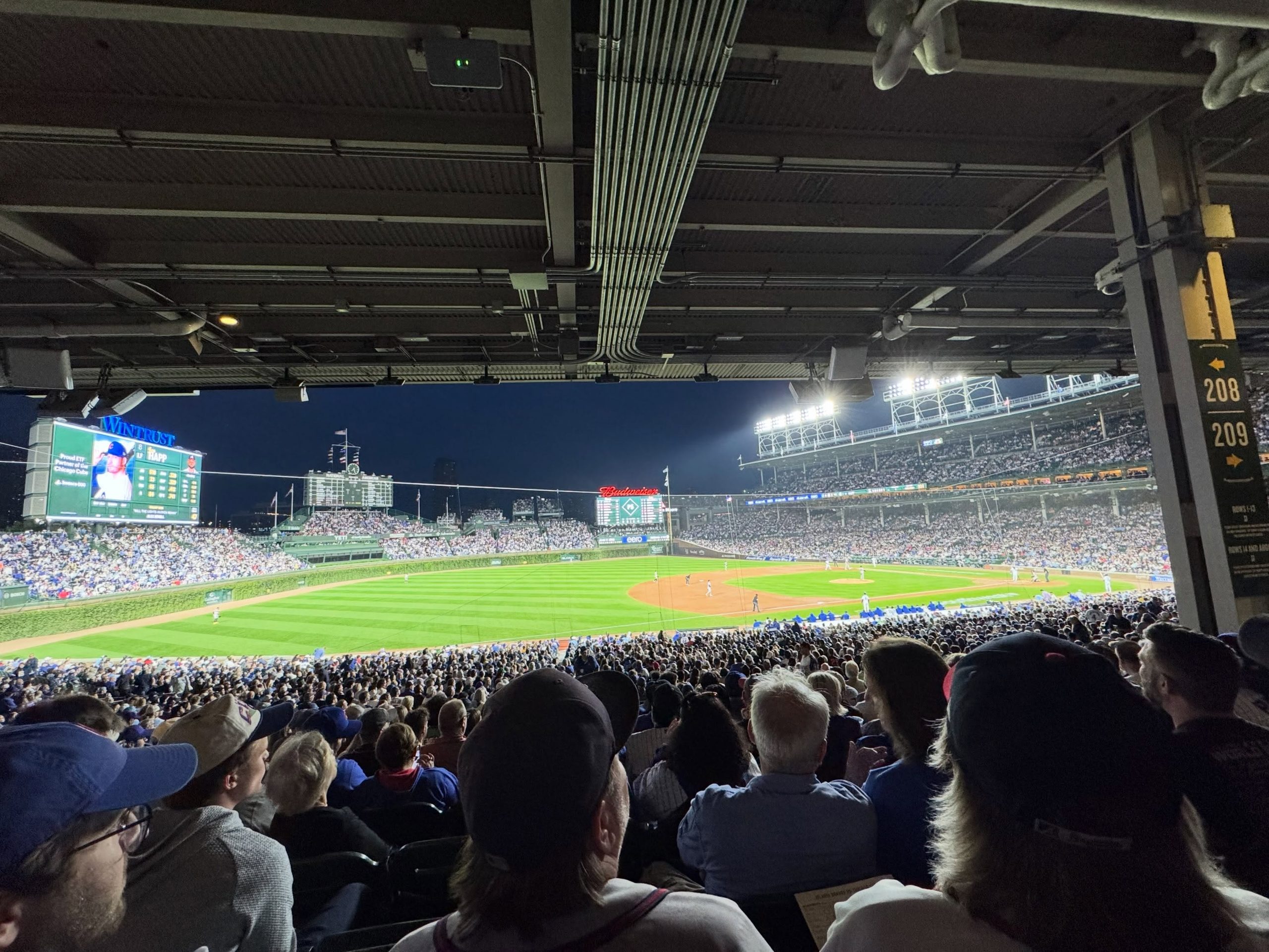 A wide view of a night baseball game at Wrigley Field, taken from under the overhang of the grandstands. Fans fill the lower-level seats in the foreground, with the brightly lit green outfield and infield visible ahead. The large left-field Jumbotron displays player stats, while the historic manual scoreboard and outfield bleachers are visible in the background under the dark evening sky.