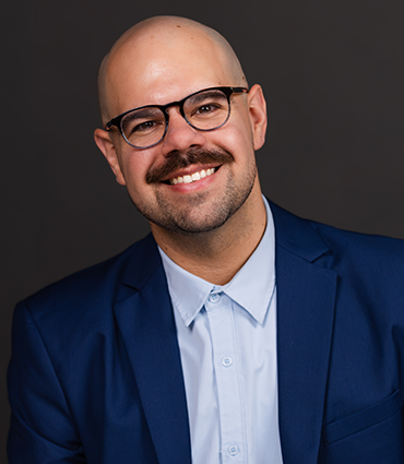 Headshot of a bald man with a mustache and goatee, wearing black round glasses, a light blue button-down shirt, and a royal blue blazer against a dark gray background.