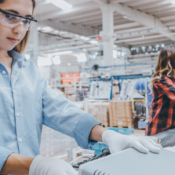 A woman wearing safety glasses and protective gloves works at an assembly station on a brightly lit manufacturing facility floor.