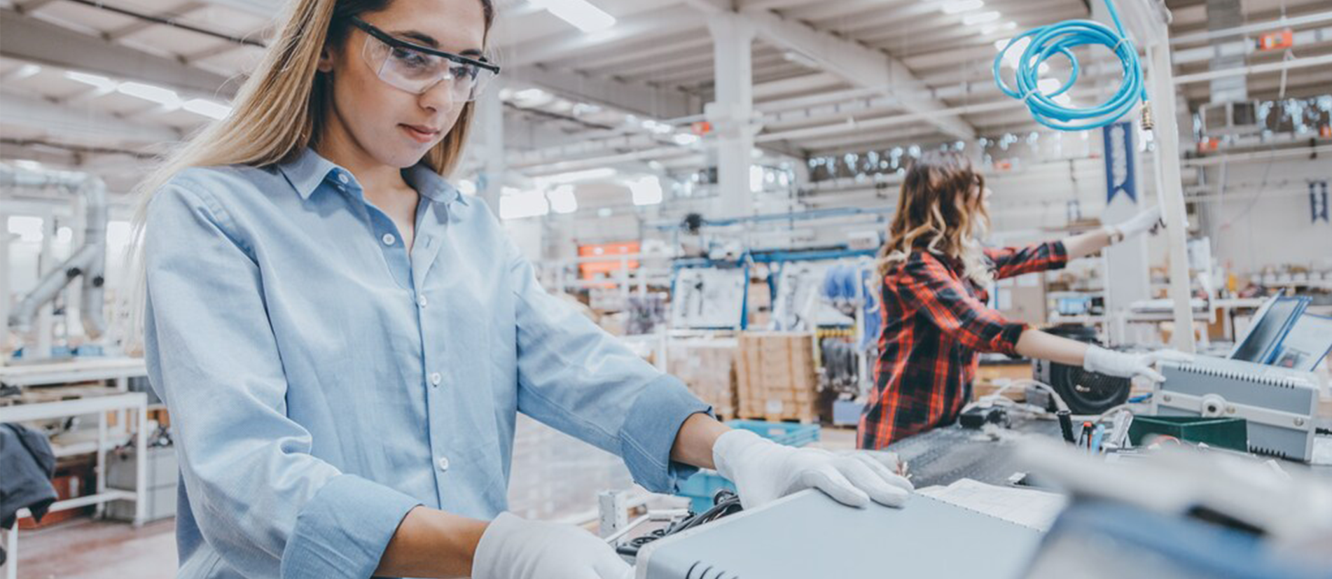 A woman wearing safety glasses and protective gloves works at an assembly station on a brightly lit manufacturing facility floor.